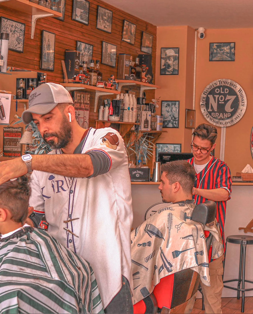 Two barbers mid-cut, one in a striped shirt working on a client under the Old No. 7 wall sign
