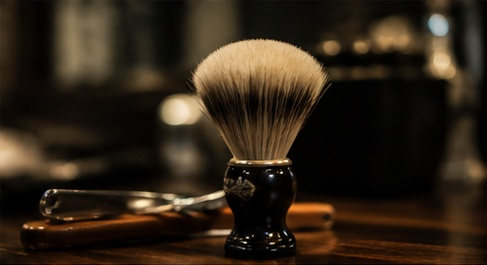A badger shaving brush standing next to an open straight razor on the wooden counter