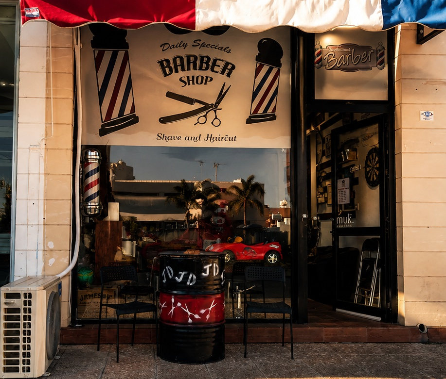 The shopfront under a red white and blue awning, painted Barber Shop sign above the window and a JD-stencilled oil drum on the pavement